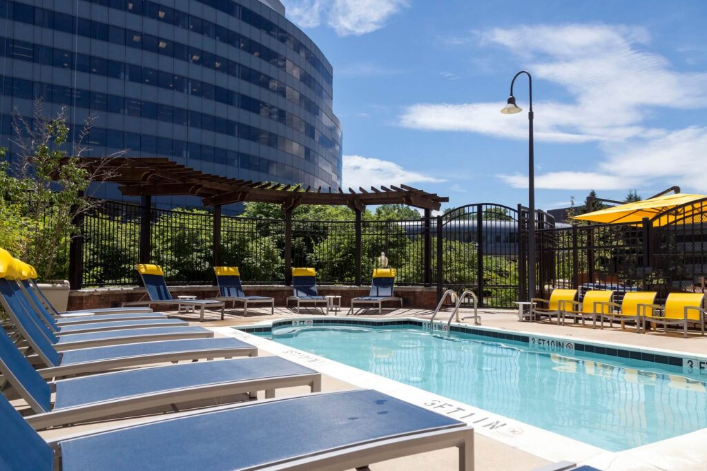 Outdoor pool area with blue lounge chairs, yellow towels, and umbrellas. A modern glass office building and greenery are visible in the background under a sunny, blue sky.