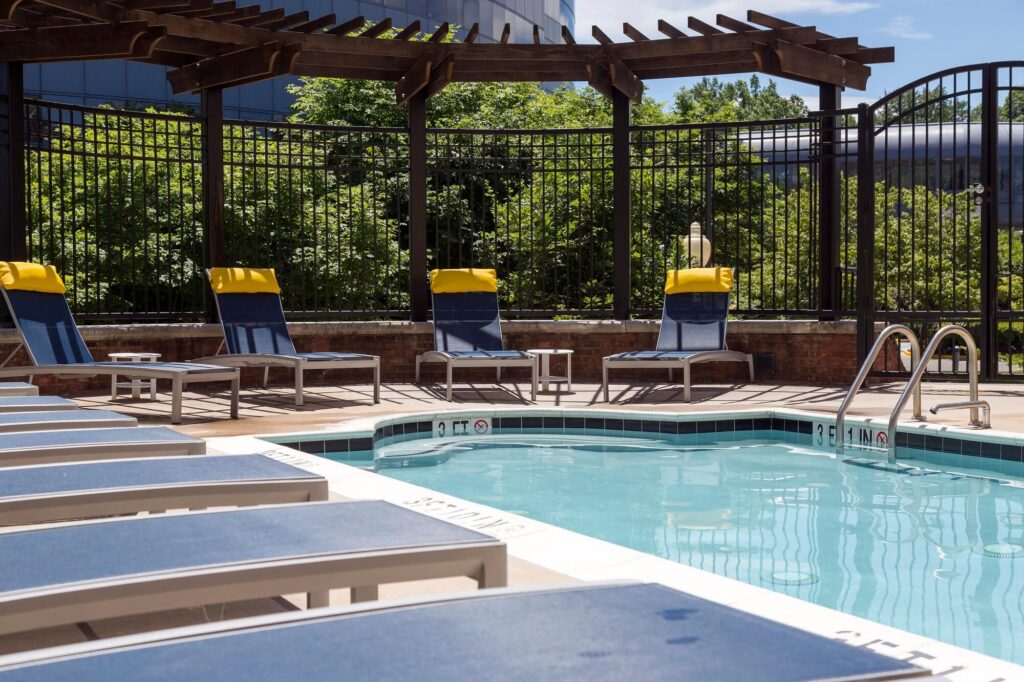 Several blue lounge chairs with yellow towels are lined up by a pool, surrounded by a black metal fence and greenery. A wooden pergola provides partial shade. The pool has clear water and pool ladders.