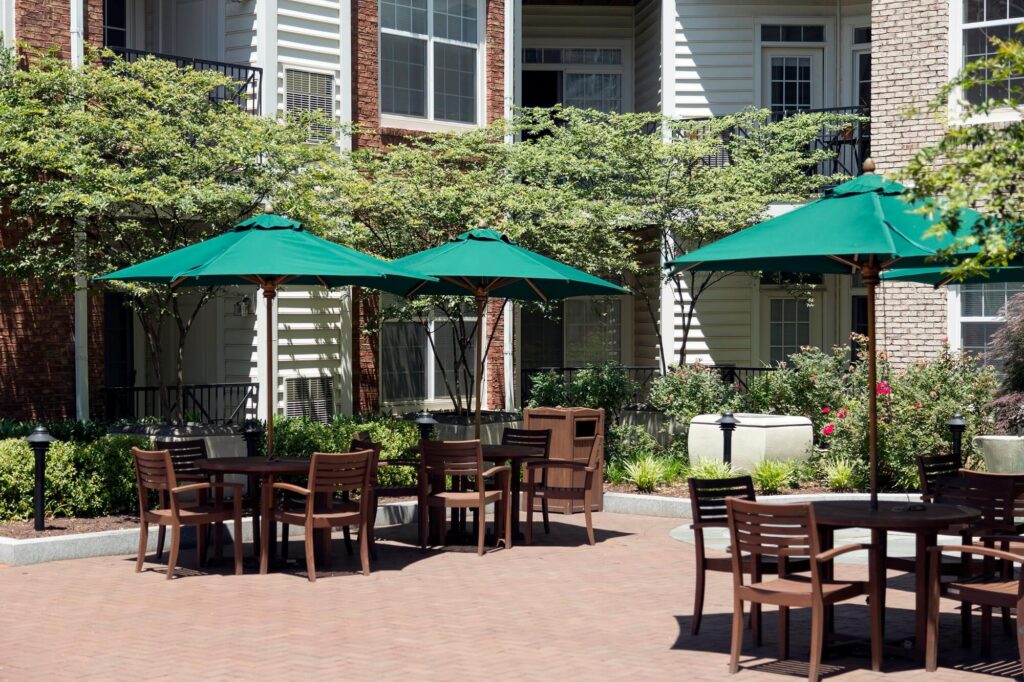 Outdoor patio area with wooden tables and chairs under green umbrellas, surrounded by plants and flowers, situated in front of a modern apartment building.