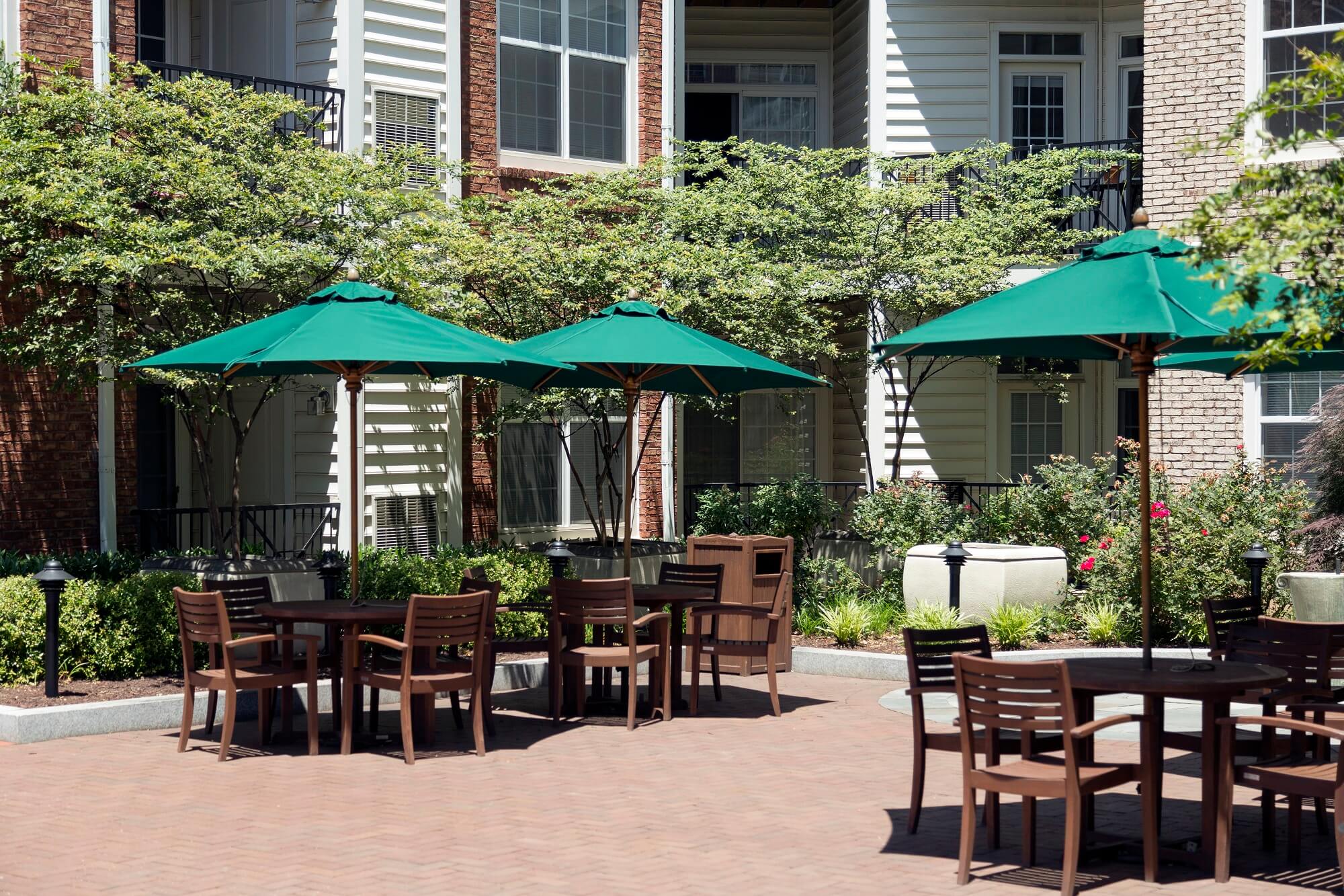 Outdoor patio area with wooden tables and chairs under green umbrellas, surrounded by plants and flowers, situated in front of a modern apartment building.