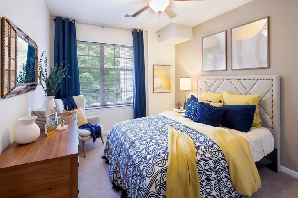 A modern bedroom with a patterned blue and white bedspread, yellow pillows and throw, wooden dresser, armchair, large window with blue curtains, and abstract art on beige walls.