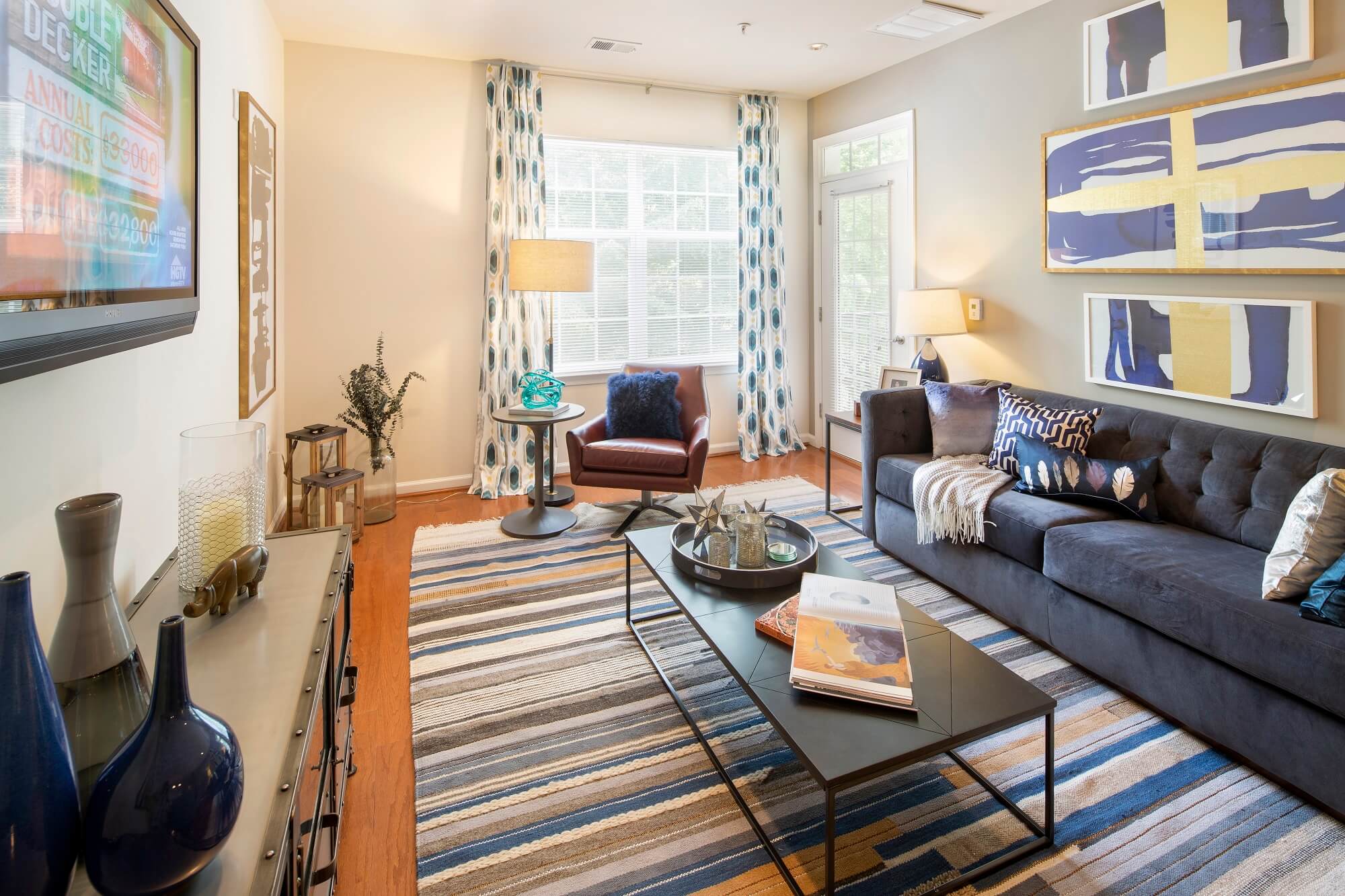 Bright living room with a striped rug, dark sofa, glass coffee table, abstract wall art, large window with patterned curtains, and various decor accents.