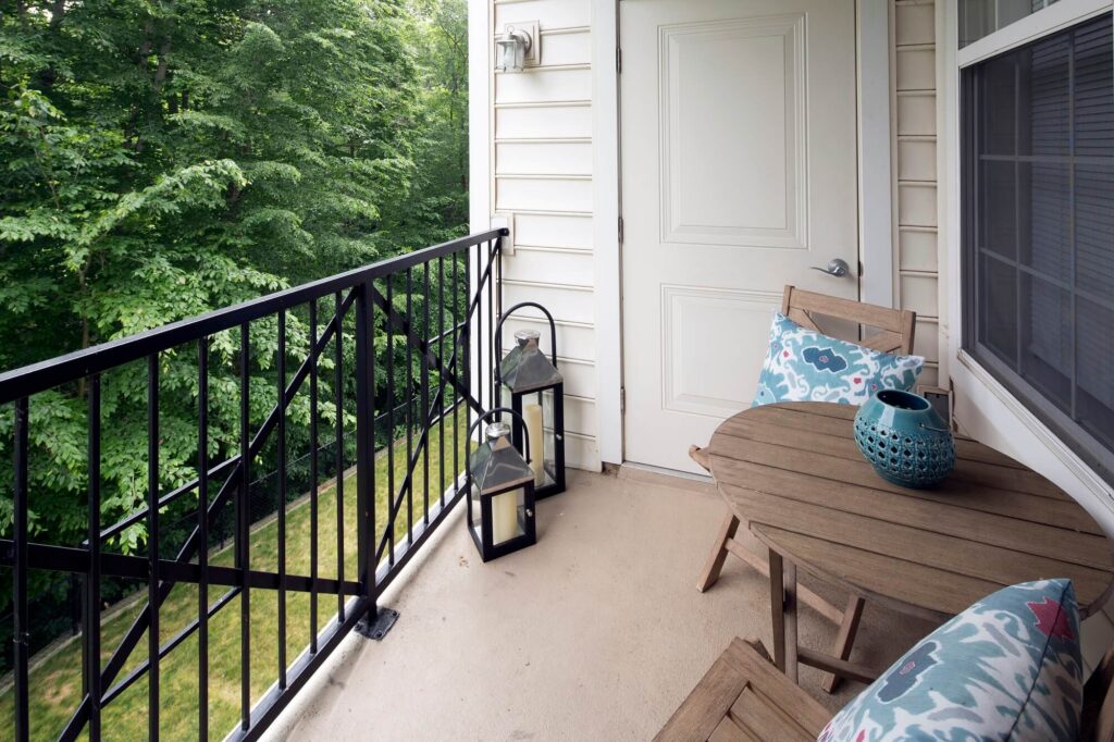 A small balcony with a wooden table and chairs, decorative cushions, two lanterns, and a blue vase. The balcony overlooks green trees and grass.