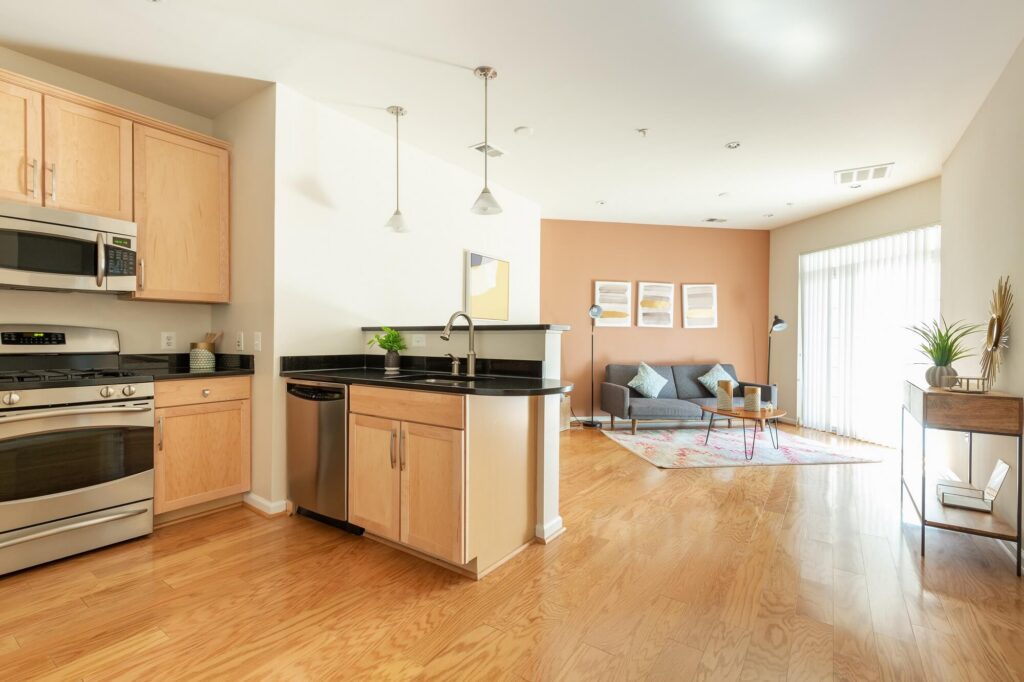 Modern open-plan kitchen with light wood cabinets and stainless steel appliances, opening to a bright living area with a sofa, art on the wall, rug, table, and large window with sheer curtains letting in natural light.