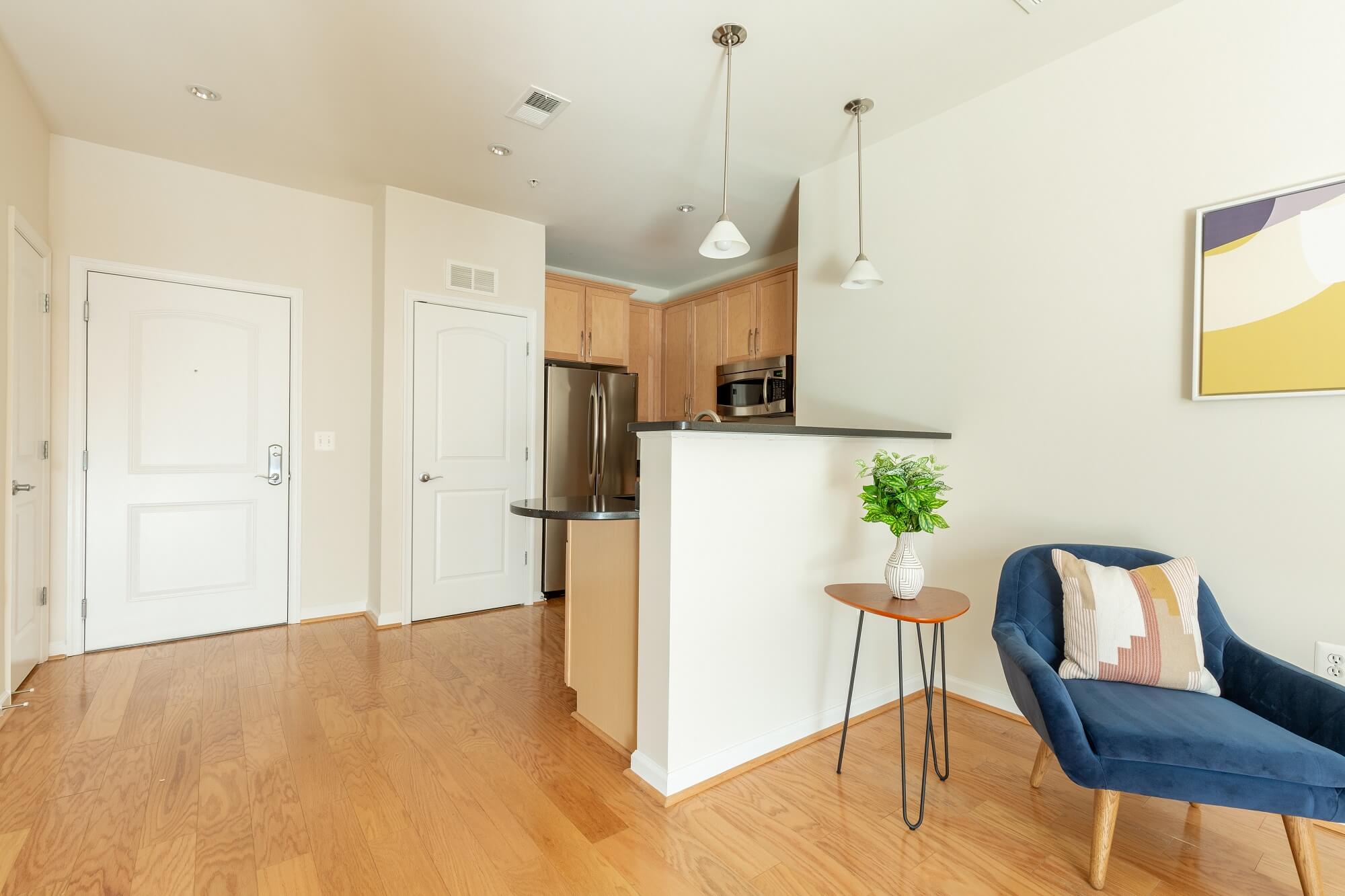 A modern apartment interior with wood floors, a small kitchen, a round table with a plant, a blue chair with a pillow, and abstract art on a cream-colored wall.