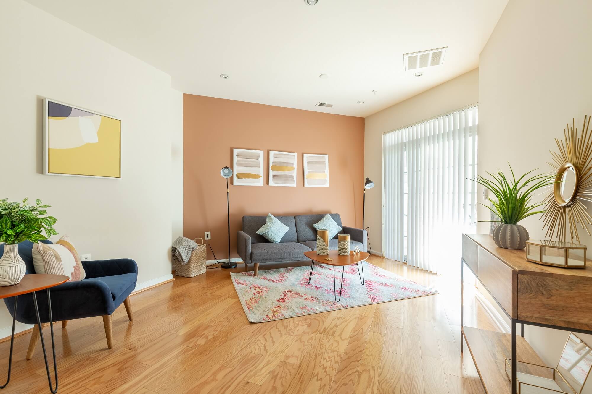 Bright living room with wood floors, a gray sofa, blue armchair, patterned rug, coffee table, abstract art on a peach accent wall, and console table with plants and decor near large windows with blinds.