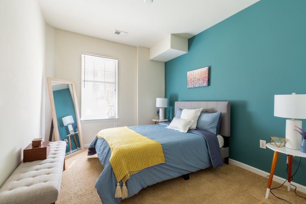 A modern bedroom with a blue accent wall, bed with blue bedding and yellow blanket, a standing mirror, a bench, two white lamps, and a window with blinds letting in natural light.