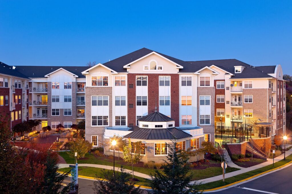 A modern, multi-story apartment building with brick and white siding, large windows, balconies, and landscaped grounds, photographed at dusk with warm interior lights visible.