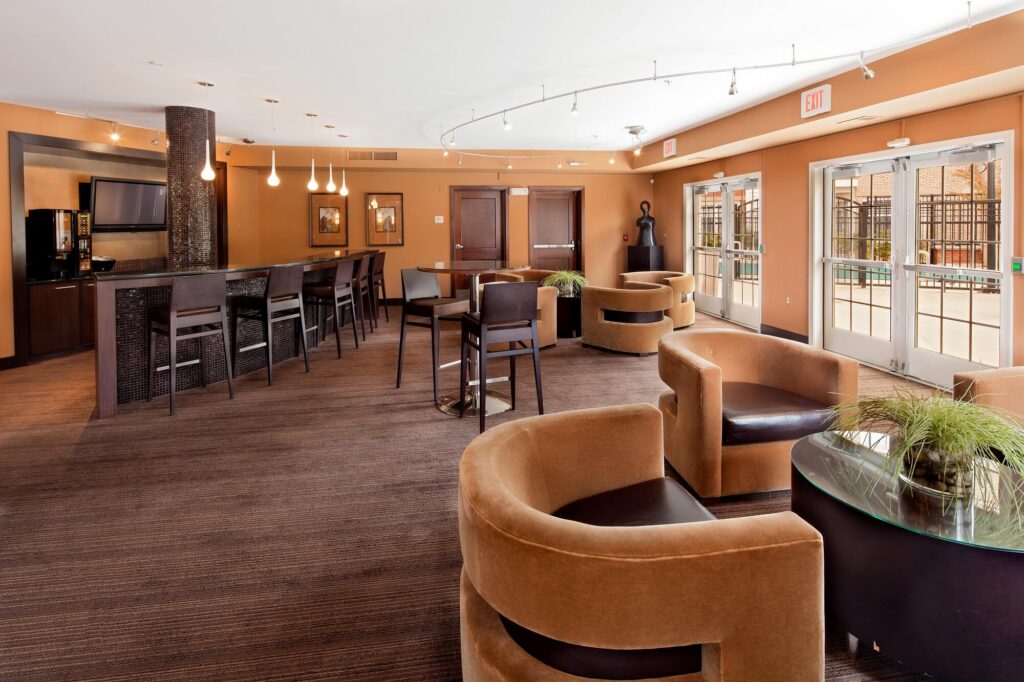 Modern hotel lounge with brown chairs, round tables, a bar area with high stools, large windows, and glass doors leading outside, illuminated by pendant and recessed lighting.